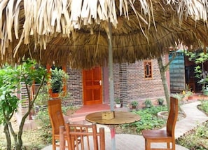 A rustic outdoor seating area with a thatched roof, wooden chairs, and a brick building with a red door in the background. - Van Long Family Homestay (Gia Vien)