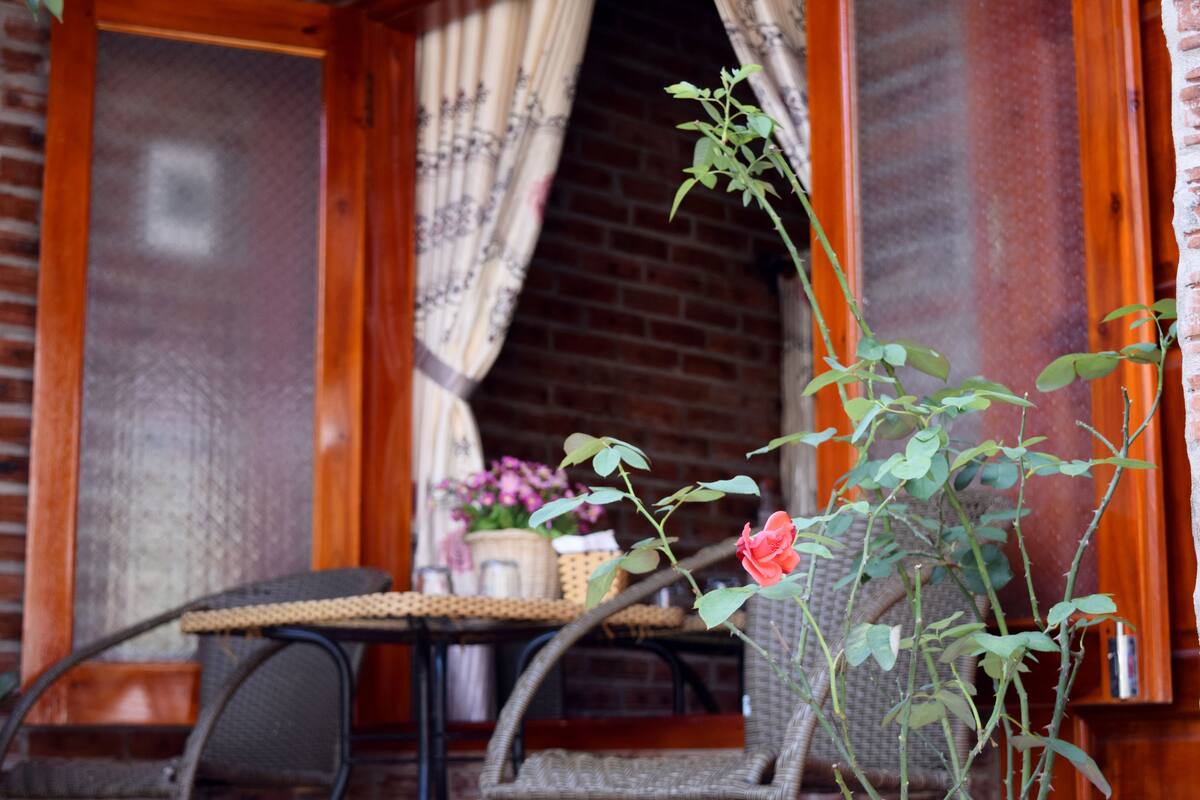 A wooden-framed window with a view of a brick wall and a wicker chair outside.