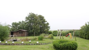 A grassy backyard with a wooden playhouse, a swing set, and a small shed. - Luxurious vacation home with a panoramic view (Bensersiel)