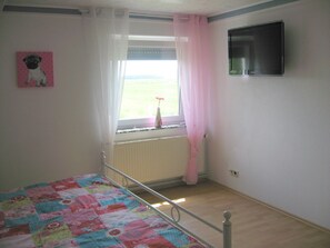 Een slaapkamer met een bed, een raam met gordijnen, een radiator en een televisie aan de muur.
