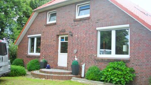 A two-story brick house with a red roof, a front door, and two windows. There is a small garden with bushes and a potted plant. - Luxurious vacation home with a panoramic view (Bensersiel)