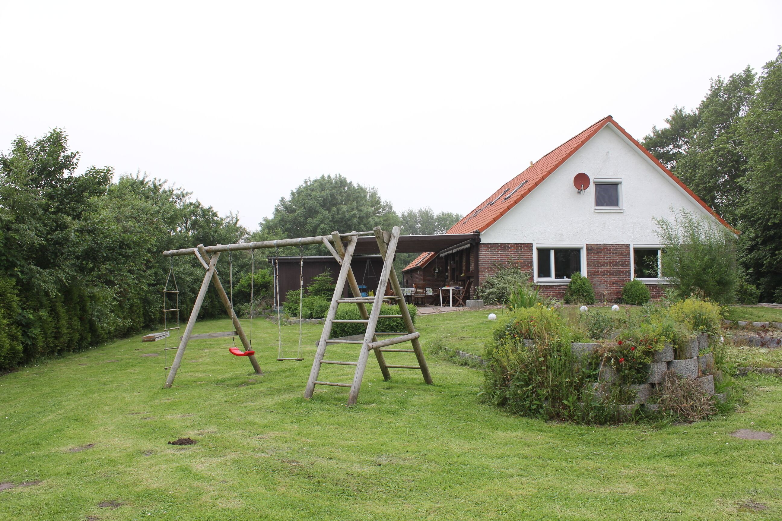Une maison avec un toit rouge et une façade en briques. Devant la maison, on trouve une balançoire et une échelle en bois appuyée contre une structure.