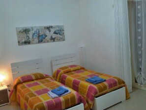 A bedroom with two beds, each with a colorful bedspread and a blue towel. There is a nightstand with a lamp, a painting on the wall, and a window with curtains. - Bed and Breakfast Ocurniciell (Naples)