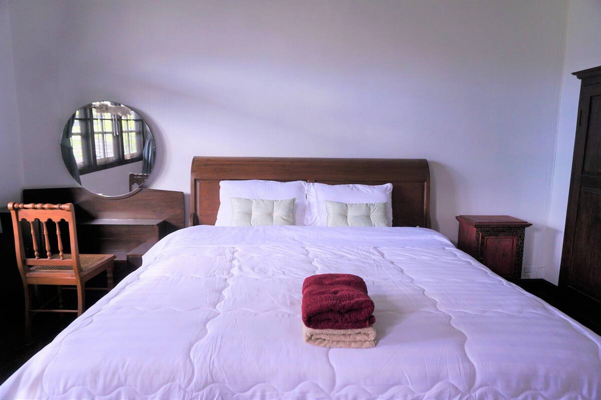 A neatly made bed with a folded red towel, a wooden chair, a small table, and a mirror on the wall.