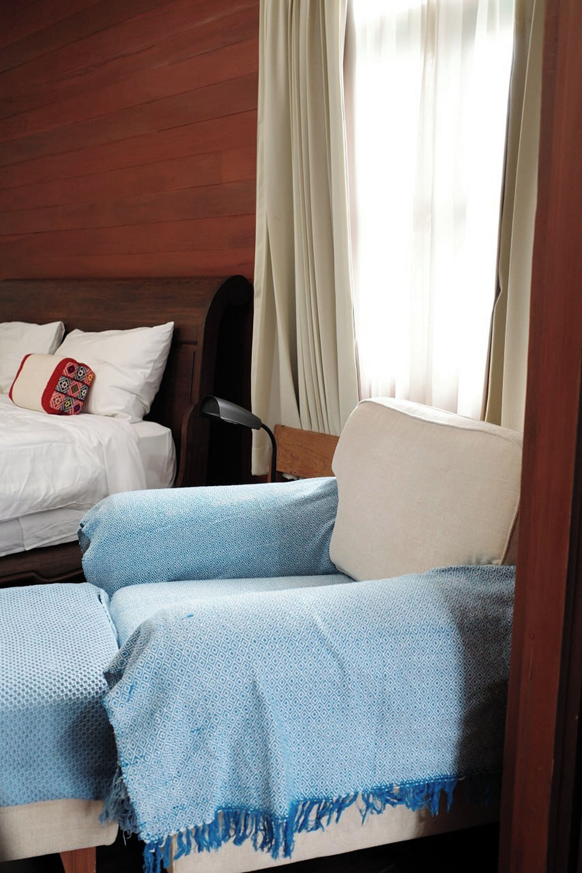 Una habitación con cabecera de madera, cama con ropa de cama blanca y almohada roja, sofá azul con estampado y una ventana con cortinas traslúcidas.
