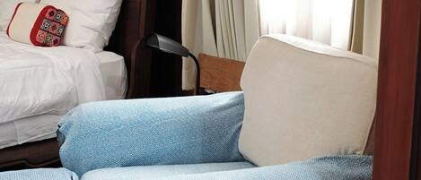 Una habitación con cabecera de madera, cama con ropa de cama blanca y almohada roja, sofá azul con estampado y una ventana con cortinas traslúcidas.
