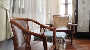 Una habitación con una silla de madera, una mesa con motivos decorativos, un armario de madera y cortinas de encaje.