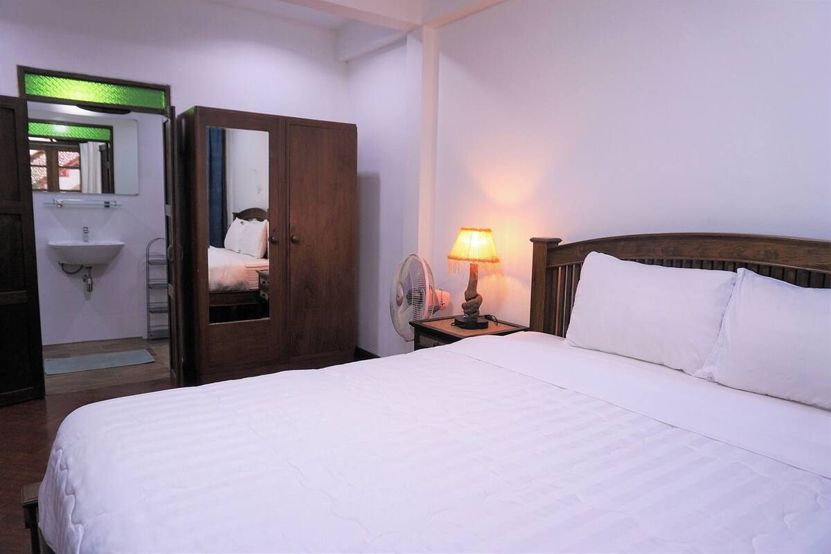 A hotel room with a bed, a wooden wardrobe, a bedside table with a lamp, a fan, and a bathroom with a sink and mirror.
