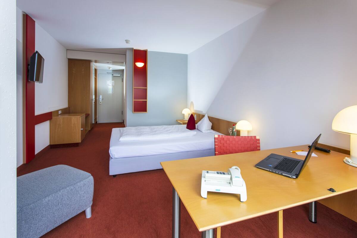Een hotelkamer met een bed, een bureau met een laptop, een nachtkastje met een lamp, een aan de muur bevestigde televisie en een rode accentmuur.