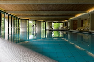 Una piscina interior con paredes de vidrio transparente, suelo de baldosas y sillones de descanso a un lado.