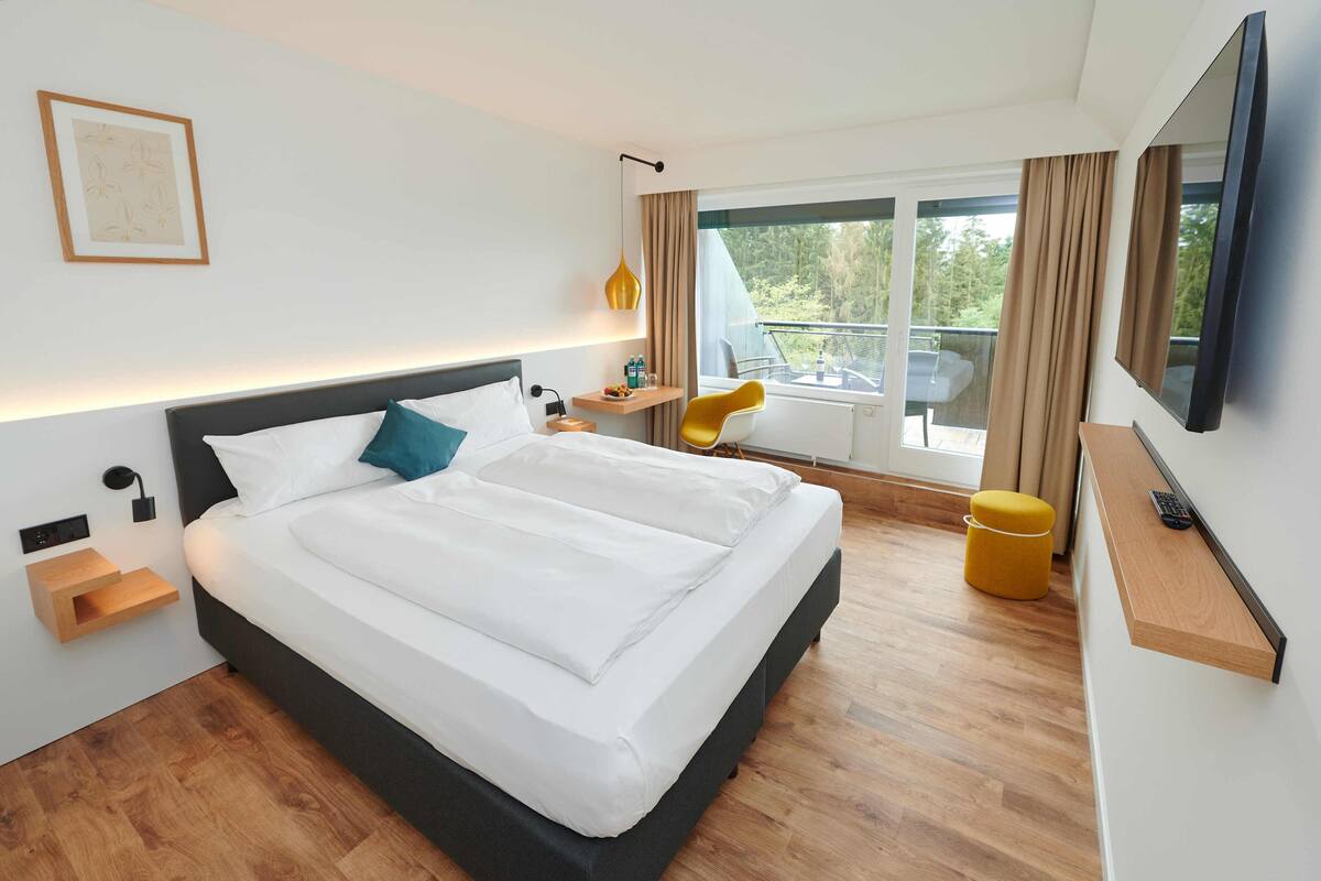 Een moderne hotelkamer met een groot bed, nachtkastjes, een televisie en een balkon met uitzicht op bomen.