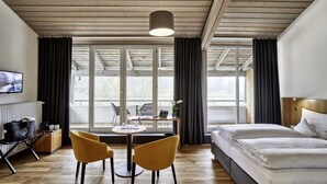 A hotel room with a bed, a desk with chairs, a television, and large windows.