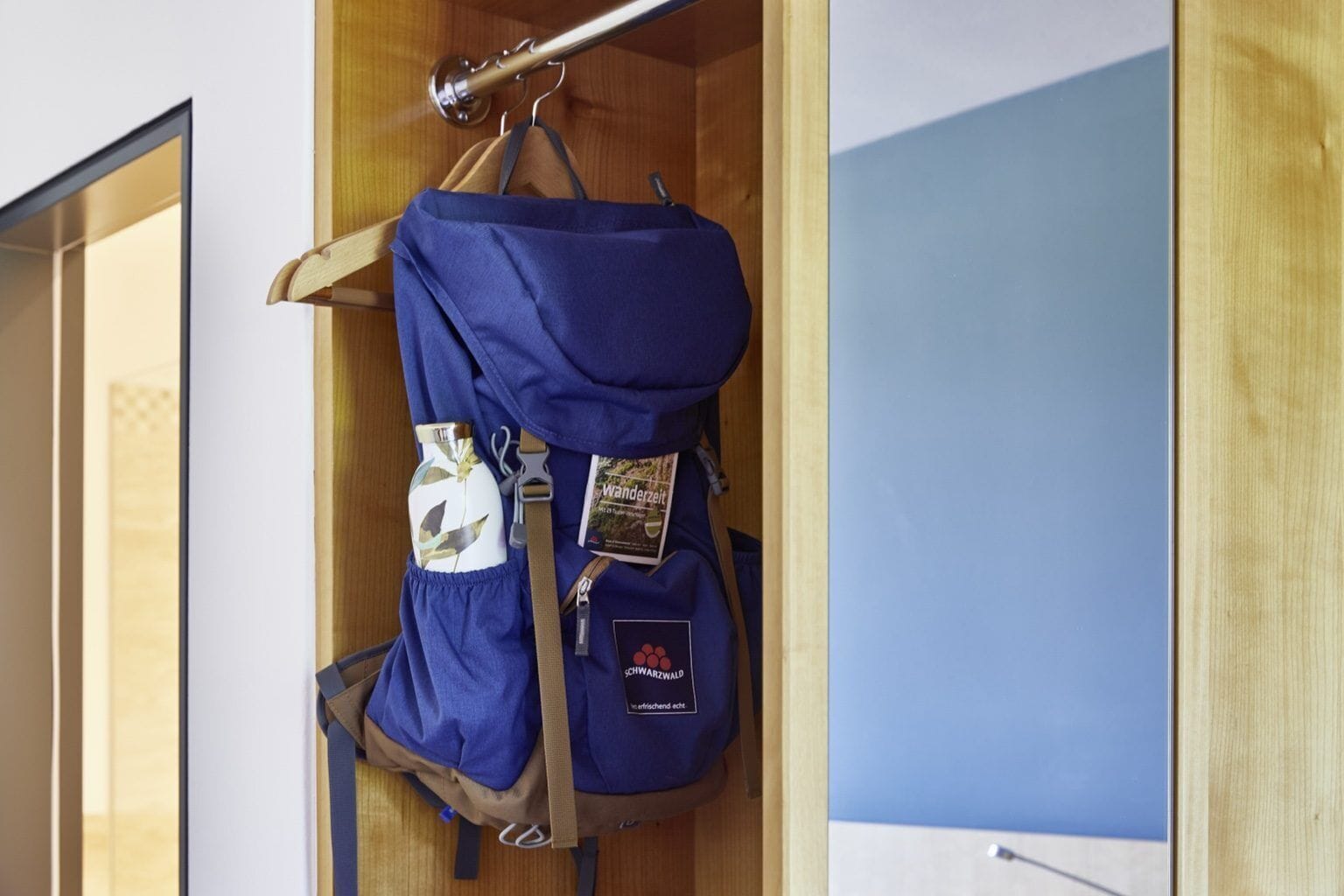 Un sac à dos bleu est accroché à un crochet métallique dans un placard en bois, sur un mur bleu.