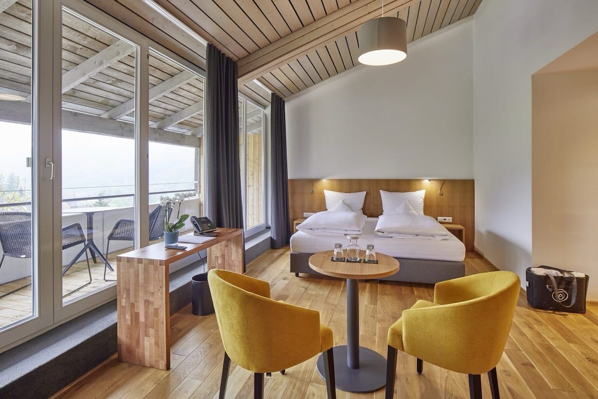 Une chambre d’hôtel avec un lit, un bureau, deux chaises, une table ronde et une vue dégagée sur l’extérieur grâce à de grandes fenêtres.