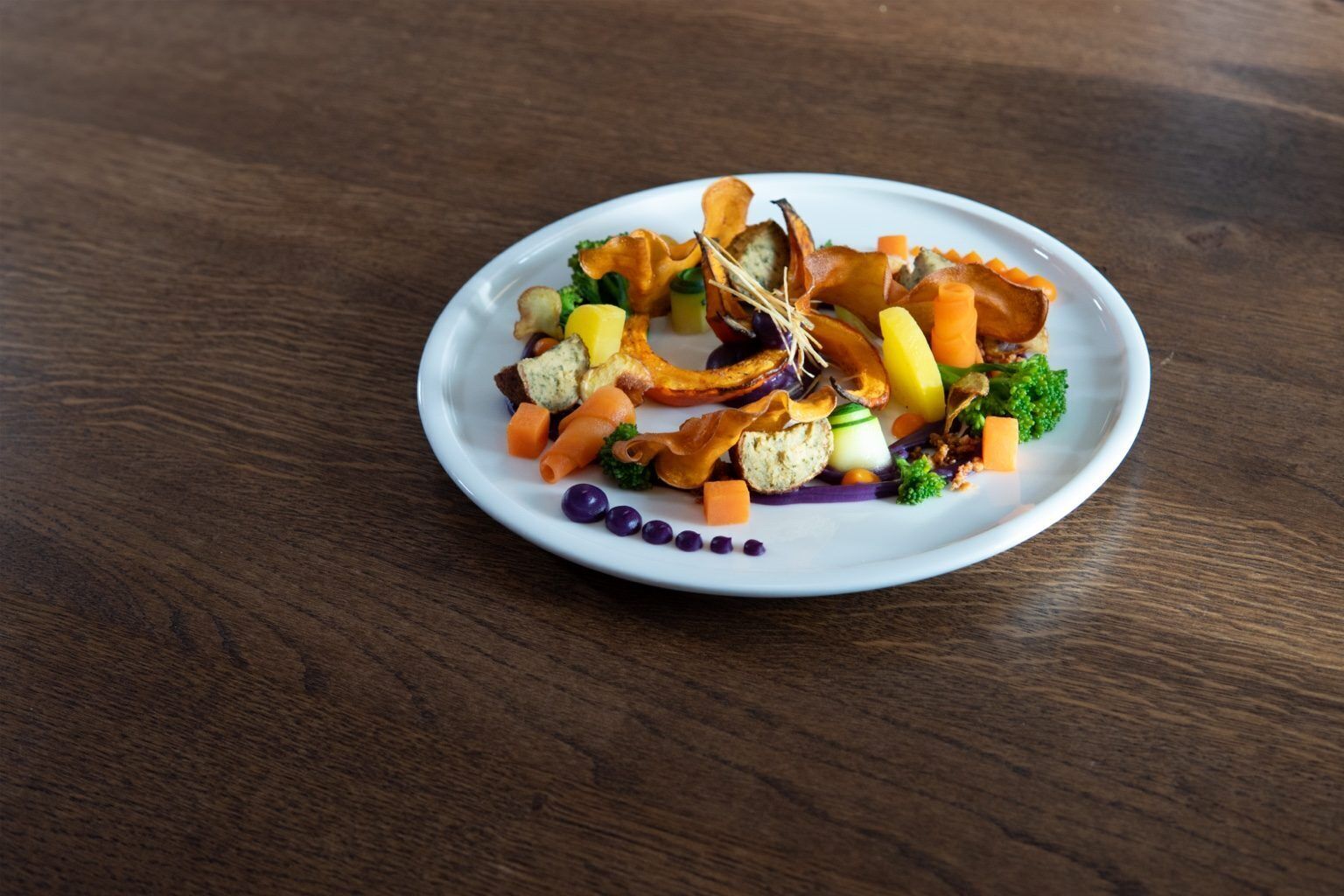 A white plate with various vegetables and a small piece of bread on a wooden table.