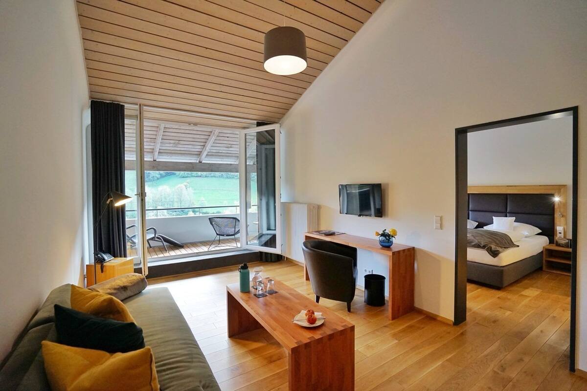 Une chambre d’hôtel moderne avec un plafond en bois, une grande fenêtre offrant une belle vue, un bureau avec une chaise, un lit et un canapé.