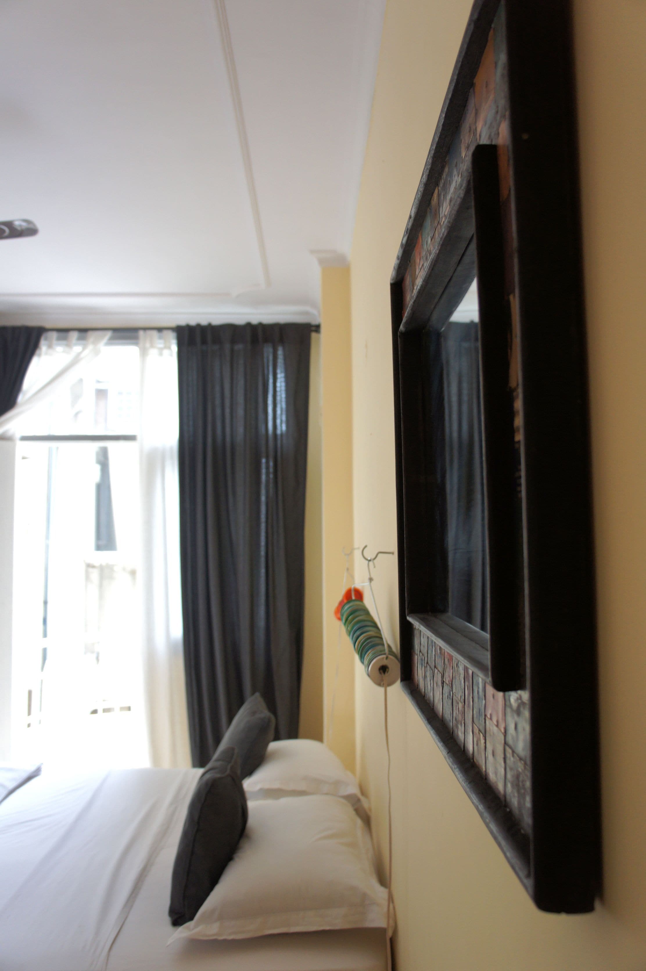 Ein Hotelzimmer mit Bett, Fenster mit Vorhängen, einem an der Wand befestigten Bilderrahmen und einem kleinen Haken, an dem ein farbiges Objekt hängt.