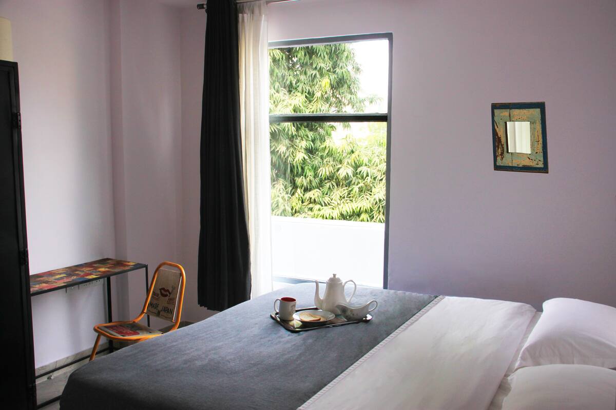 Ein ordentlich bezogenes Bett, ein Tablett mit Tee, ein Sessel und ein Fenster mit Blick auf Grünflächen.
