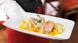 Een wit rechthoekig bord met een zeevruchtengerecht, versierd met kruiden, vastgehouden door een persoon in een uniform van rood en zwart.