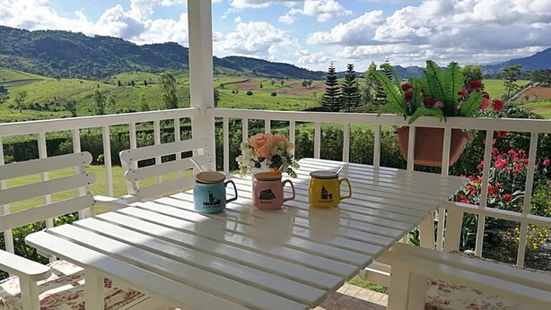 A white wooden table with three mugs on it, a bench, and a view of a green landscape with mountains in the background. - Misty English Cottage (Khao Kho)