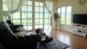 A living room with a sofa, a television, and a view of a green landscape through large windows. - Misty English Cottage (Khao Kho)