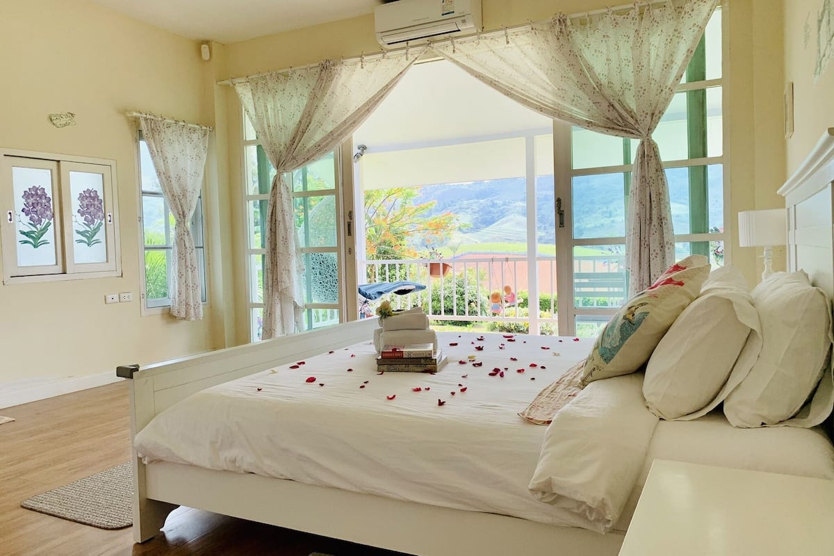 A bedroom with a bed, a bedside table, a window with curtains, and a view of the outdoors.
