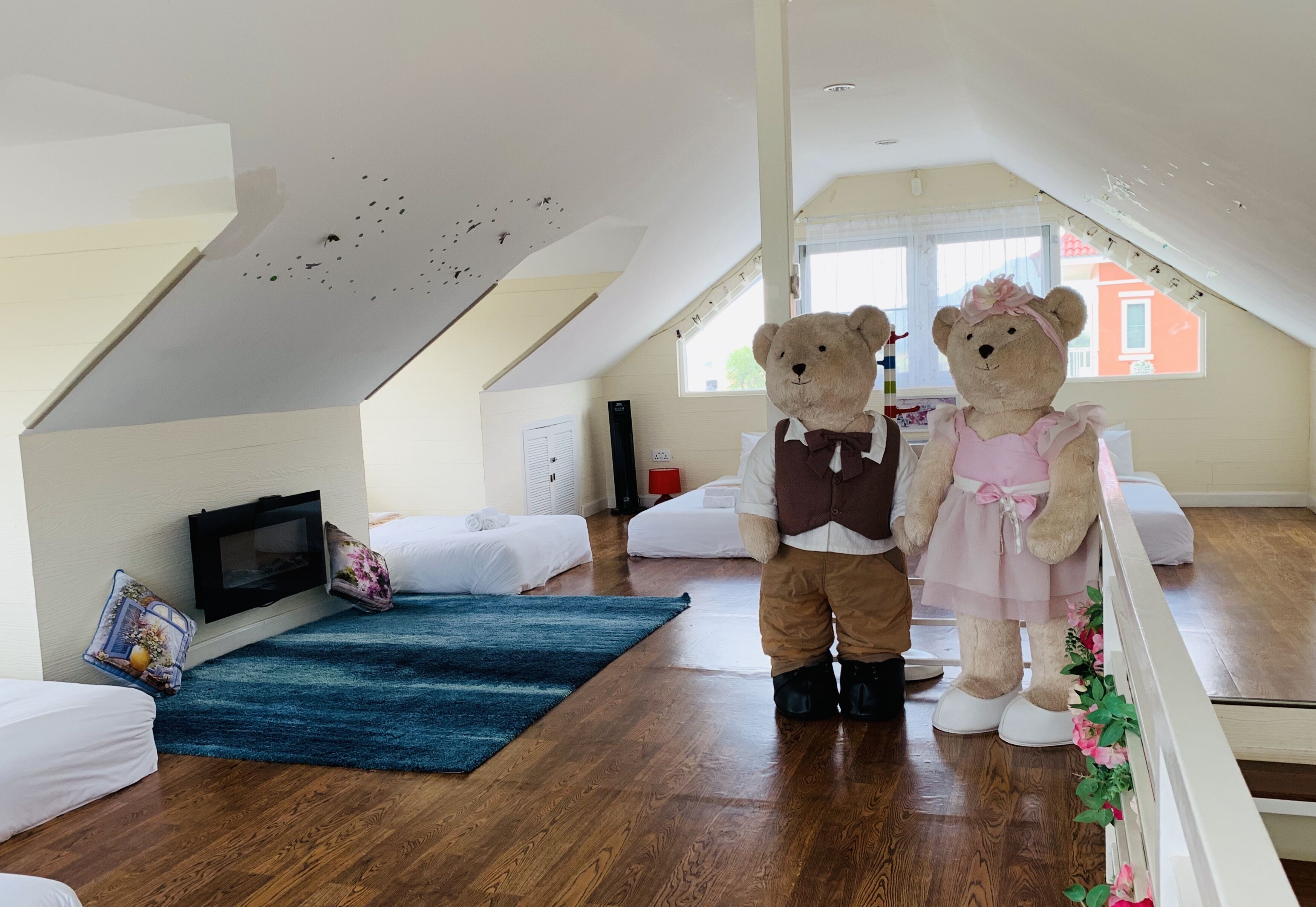 Une pièce avec un plafond en pente, une télévision, deux lits, un tapis bleu et un ours en peluche habillé d’une robe.