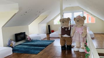 Une pièce avec un plafond en pente, une télévision, deux lits, un tapis bleu et un ours en peluche habillé d’une robe.