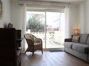 Une pièce comprenant une chaise en osier, un canapé gris et une porte-fenêtre coulissante donnant sur un espace extérieur avec une clôture blanche et des arbres.