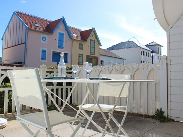 A patio table with white chairs, a bottle, and glasses, set against a white fence with houses in the background.