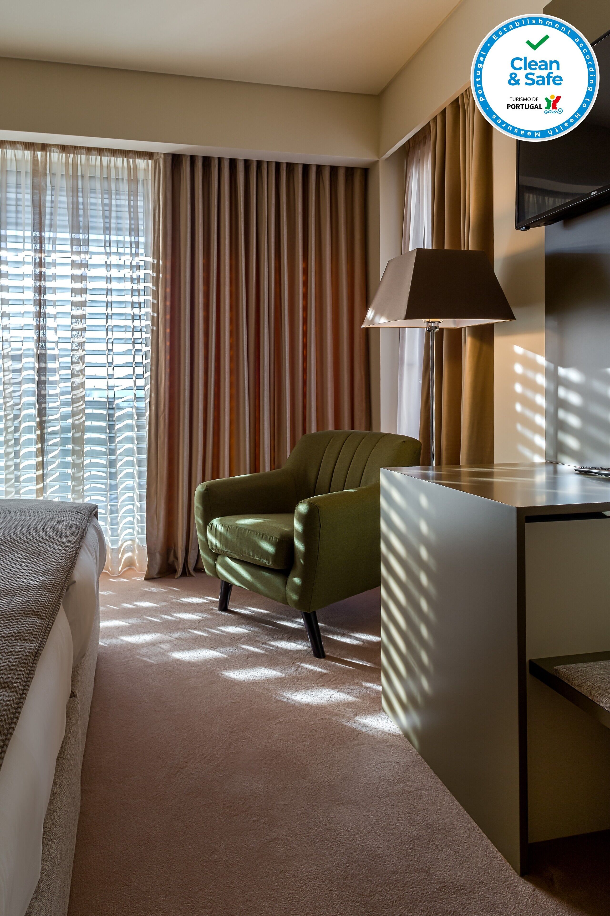 A hotel room with a bed, a green armchair, a desk, and a TV mounted on the wall.