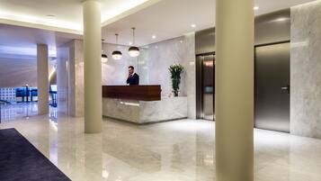 A modern hotel lobby with a reception desk, large pillars, a plant, and a visible elevator.
