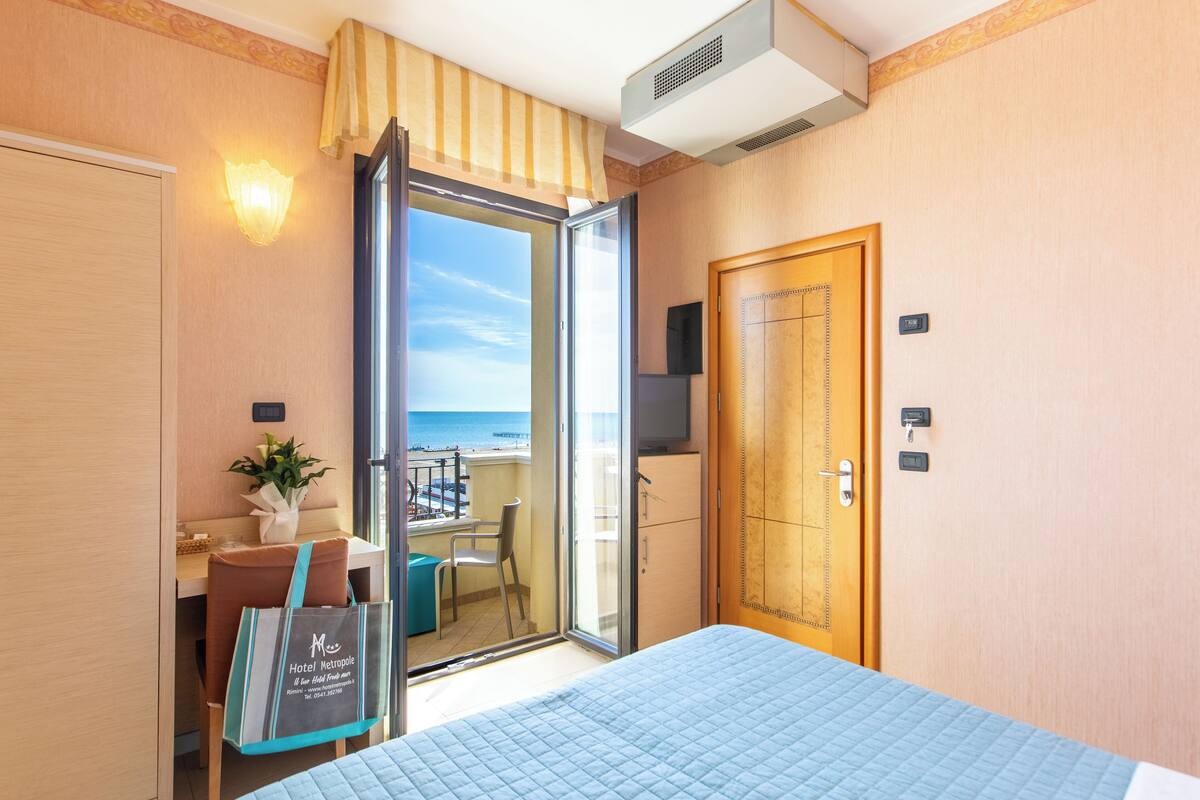 Una camera d'albergo con un letto, una scrivania con una sedia, una pianta, una porta scorrevole in vetro-camera che dà su un balcone e una televisione appesa al muro.
