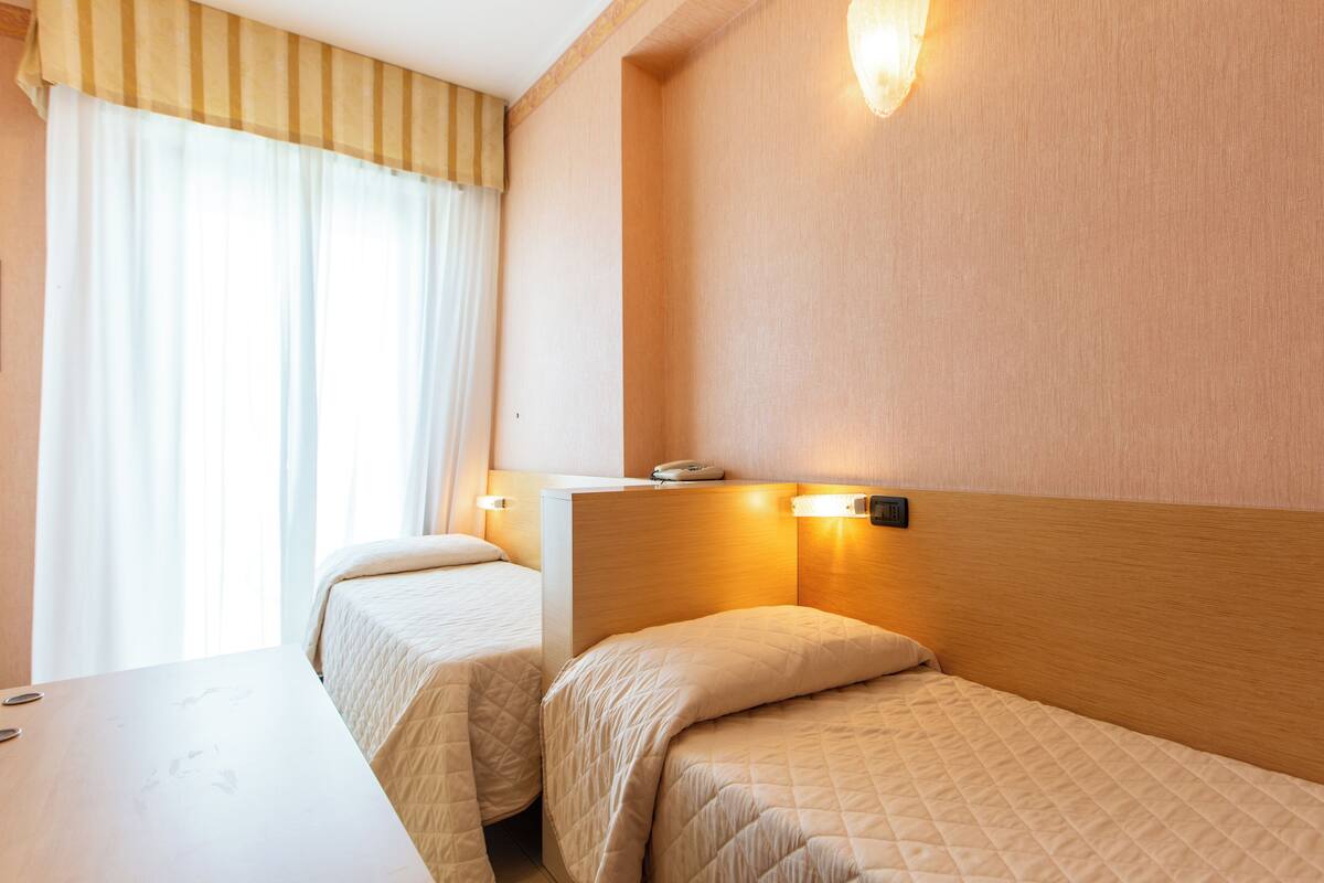 Una camera d'albergo con due letti singoli, una testiera di legno, un comodino e una finestra con tende.