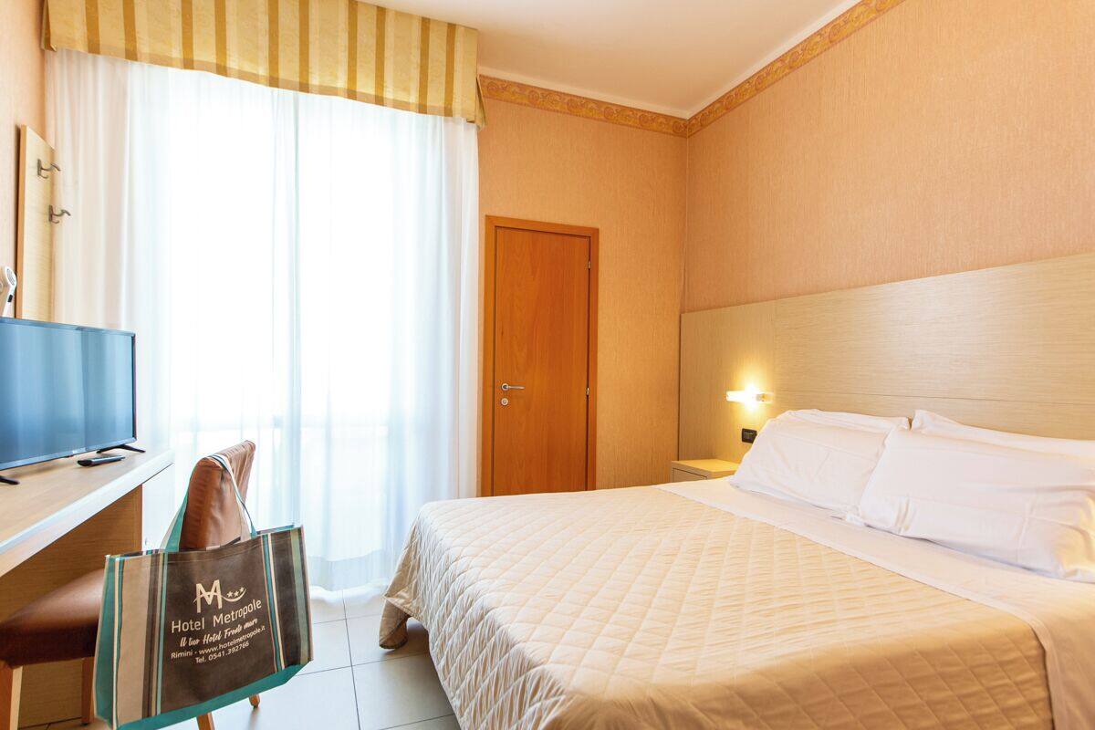 Una camera d'albergo con un letto, una scrivania con un computer, una sedia, una porta in legno e una finestra con tende.
