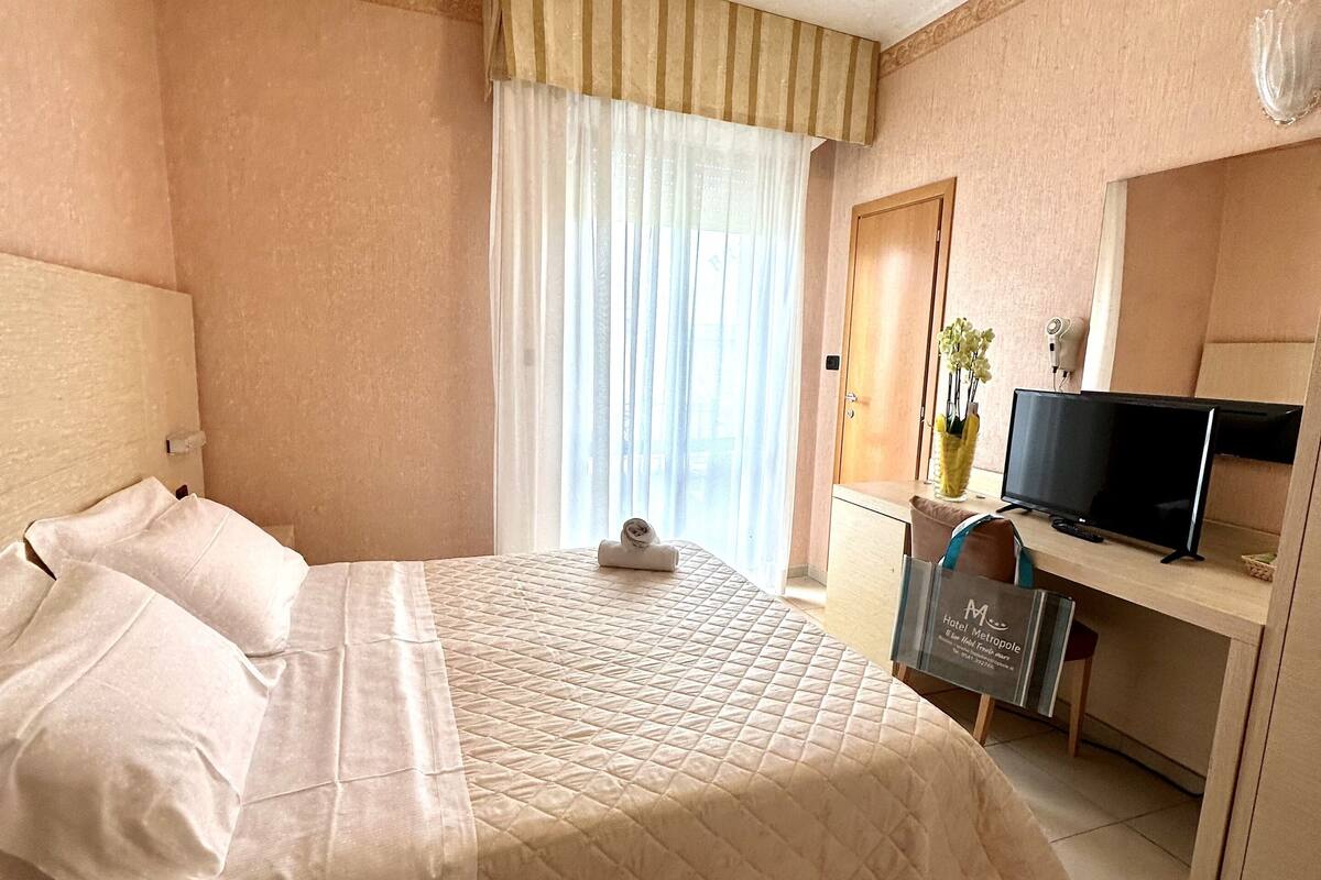 Una camera d'albergo con un letto, una scrivania con televisore, una finestra con tende e un asciugacapelli fissato al muro.