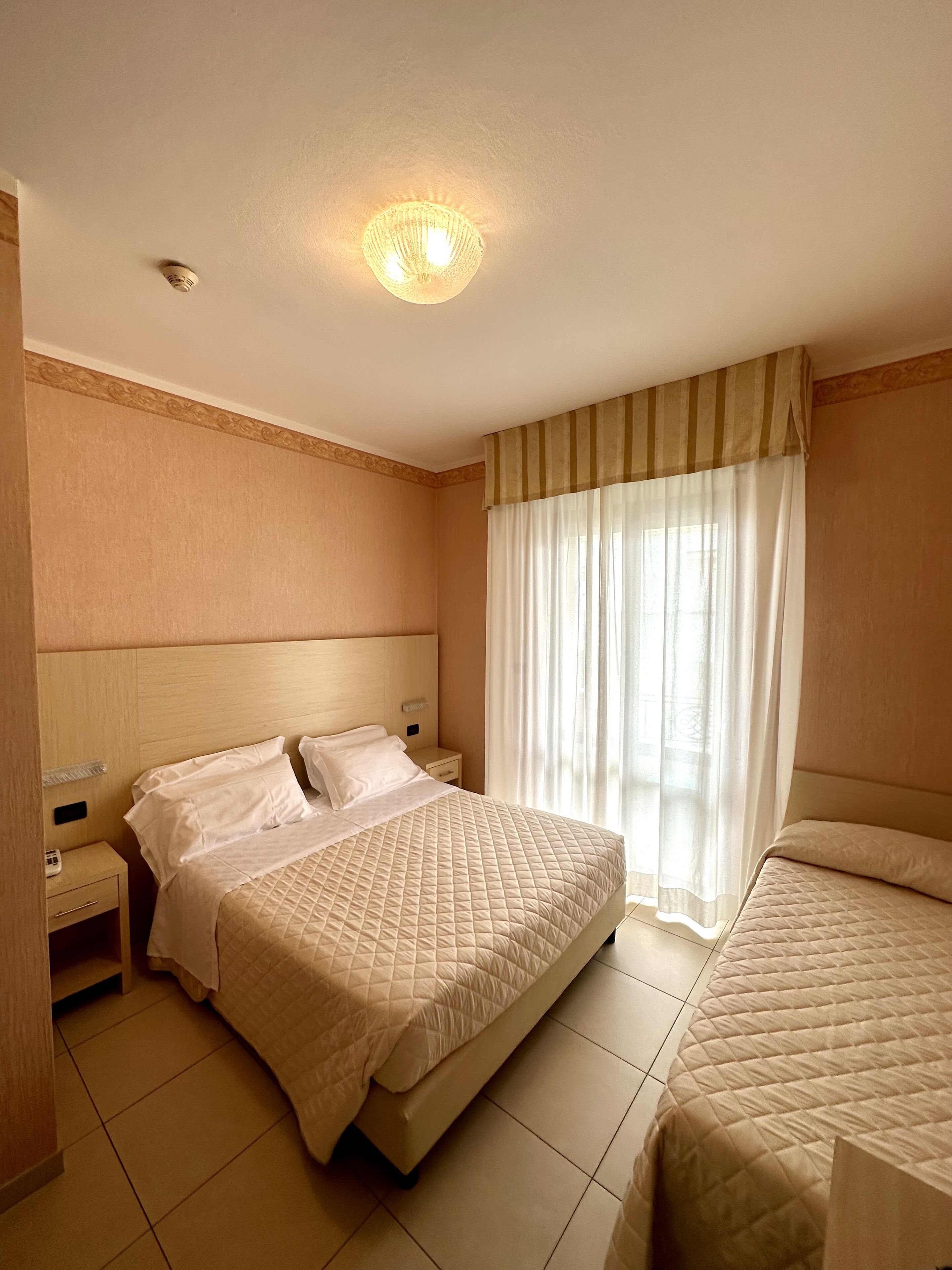 A hotel room with a large bed, a smaller bed, a nightstand, a lamp, and a window with curtains.