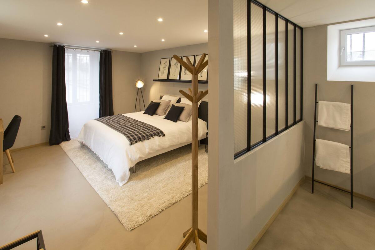 Ein modernes Schlafzimmer mit einem großen Bett, einem Nachttisch, einer Stehlampe und Blick ins Freie durch ein Fenster.