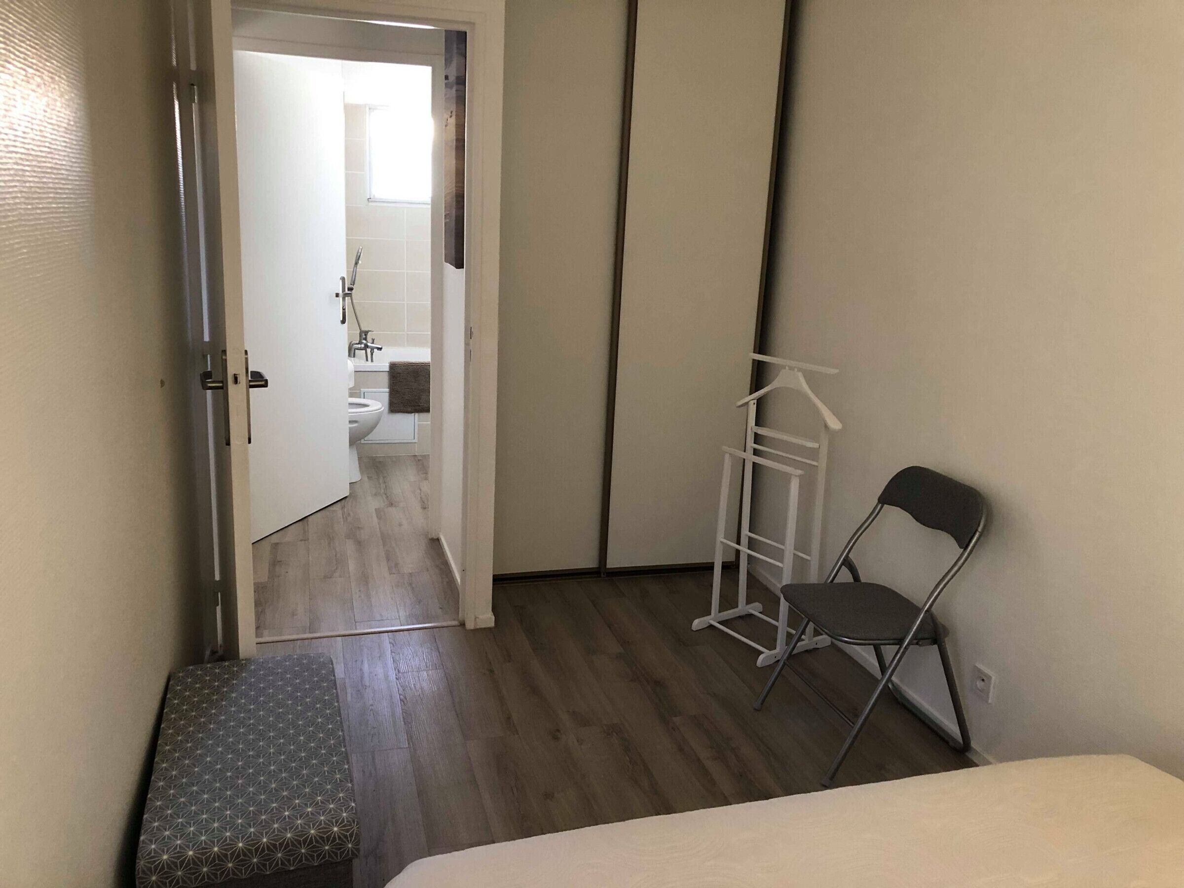 A narrow room with a bed, a bench, a hanging clothes rack, and a view into a bathroom with a toilet and sink.