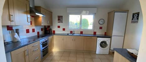 Une cuisine compacte avec des meubles en bois, un four encastré, un lave-linge et une fenêtre donnant sur un espace verdoyant.