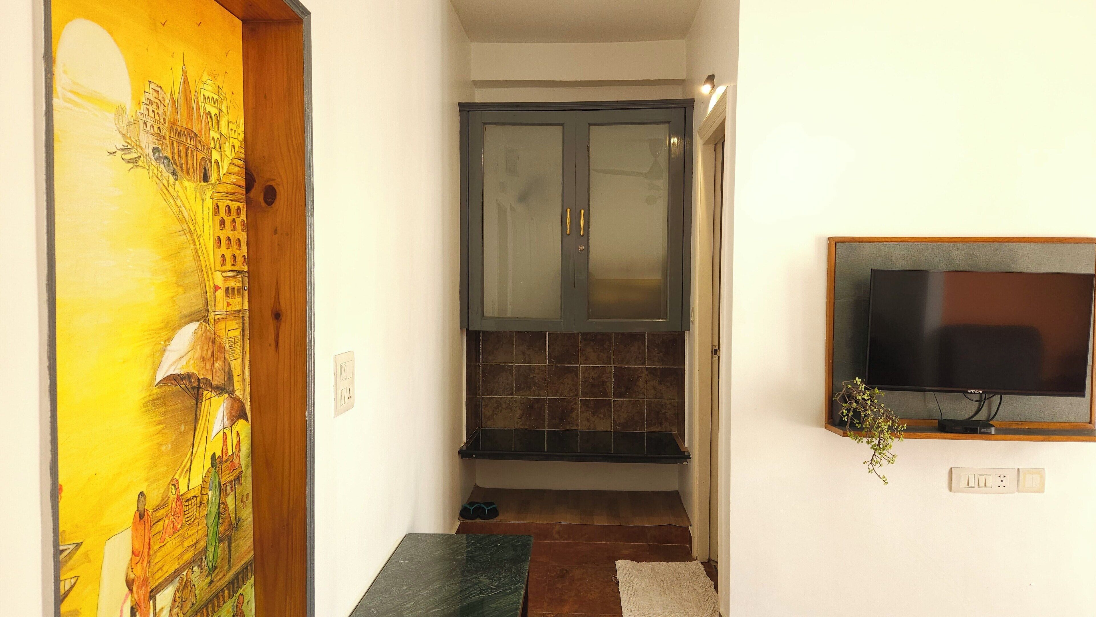 Um corredor com uma porta de madeira adornada por um mural colorido, um armário escuro com portas de vidro e uma televisão fixada na parede.