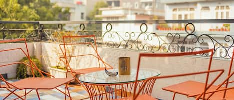 A terrace with orange metal chairs and a glass table, overlooking a cityscape with buildings and greenery.