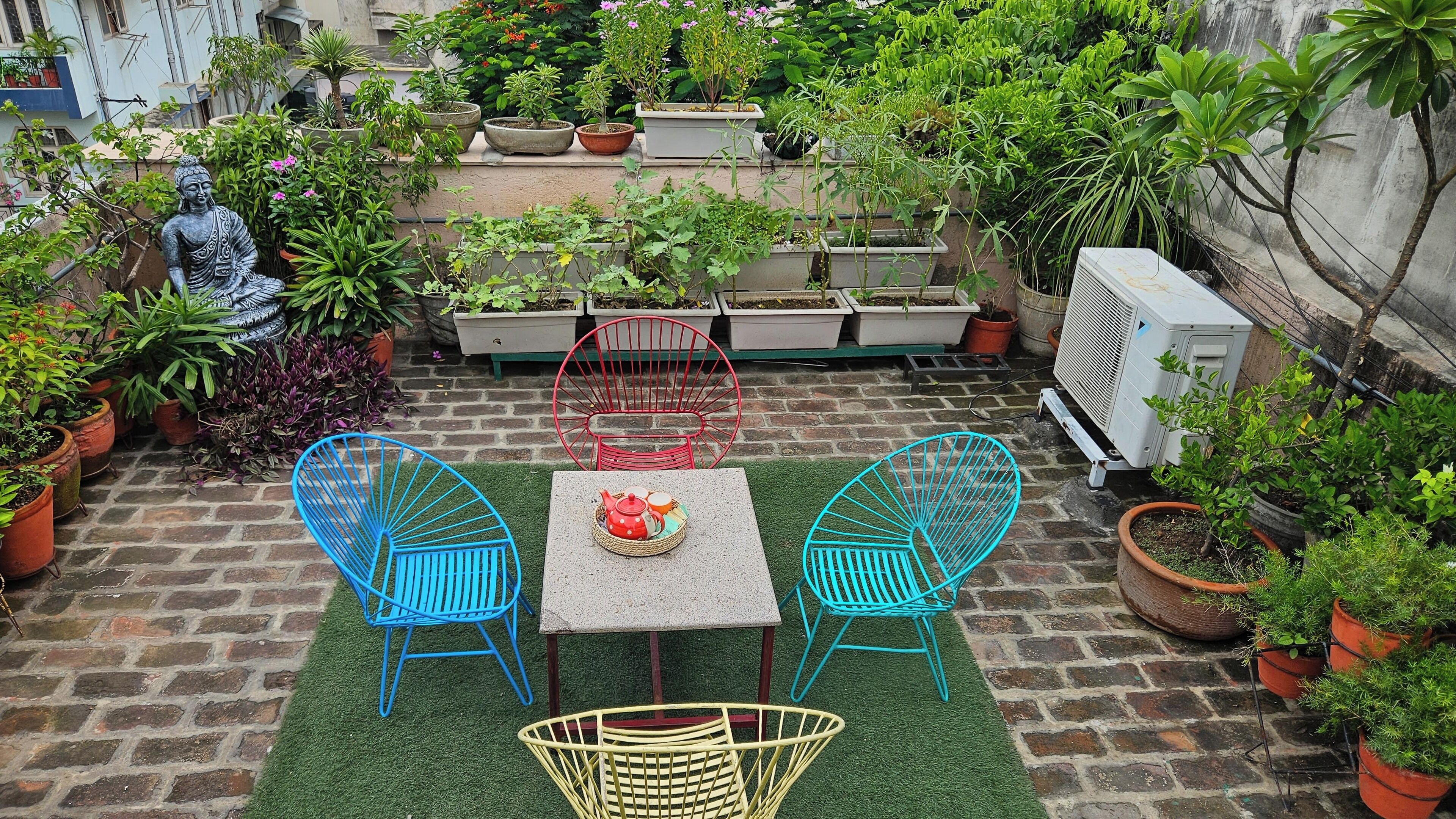 Un patio con una estatua, plantas en macetas, una mesa con sillas y un equipo de aire acondicionado.