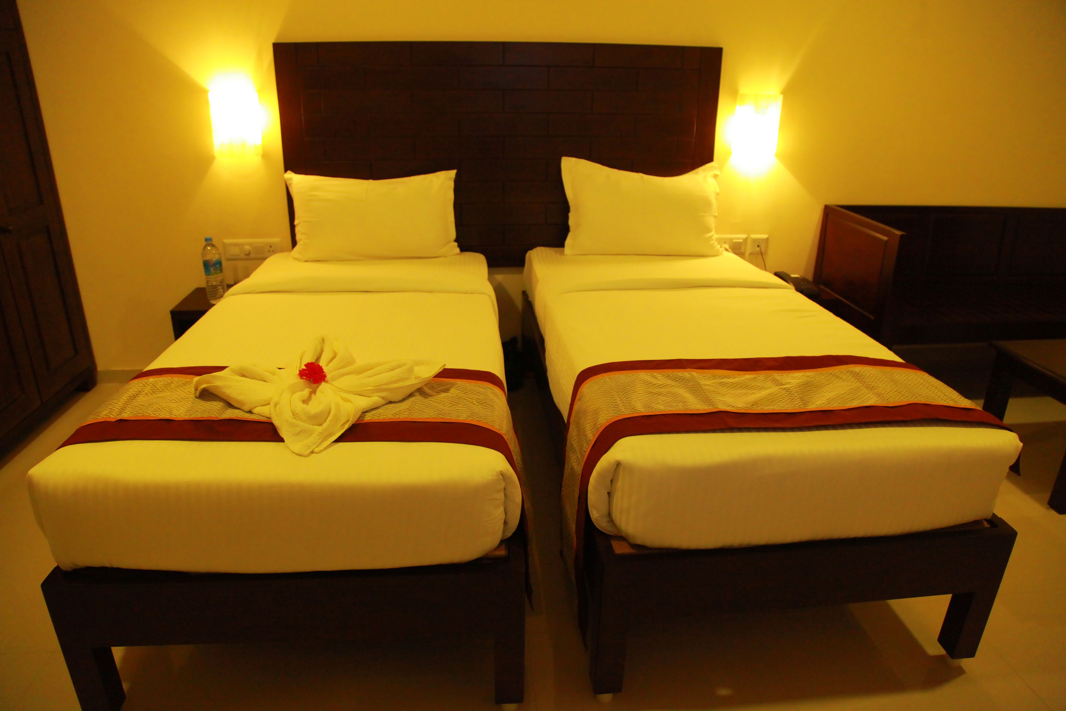 Habitación de hotel con dos camas individuales, cada una con ropa blanca y un detalle rojo. Hay dos lámparas fijadas a la pared, una a cada lado de las camas.