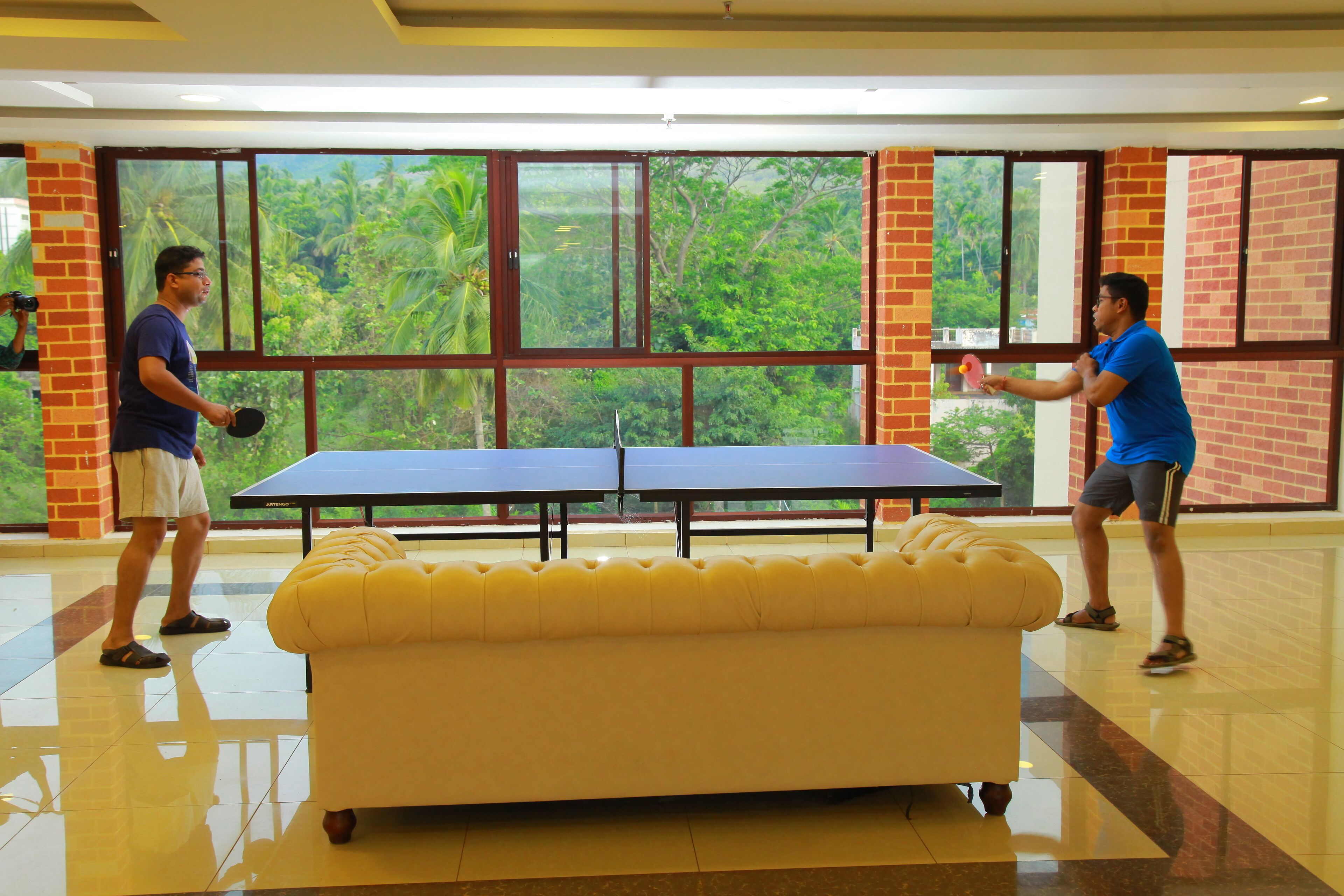 Two people playing table tennis in a room with a large window, a yellow sofa, and a brick wall.