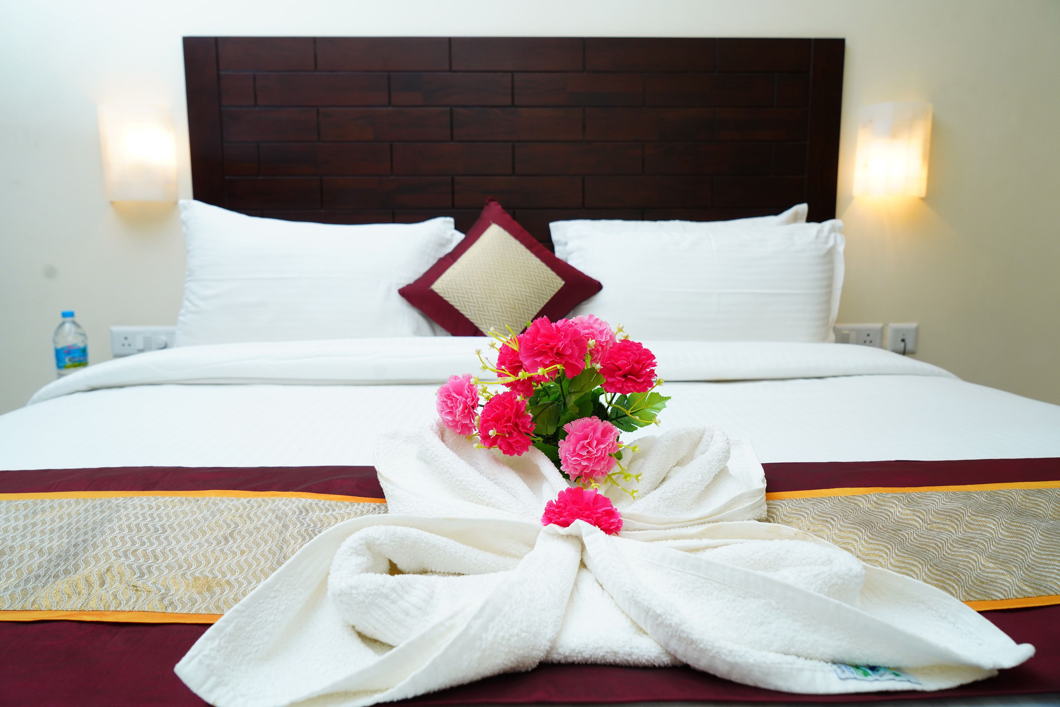 Un lit parfaitement fait, avec du linge de lit blanc, un couvre-lit rouge et doré, et un bouquet de fleurs roses.