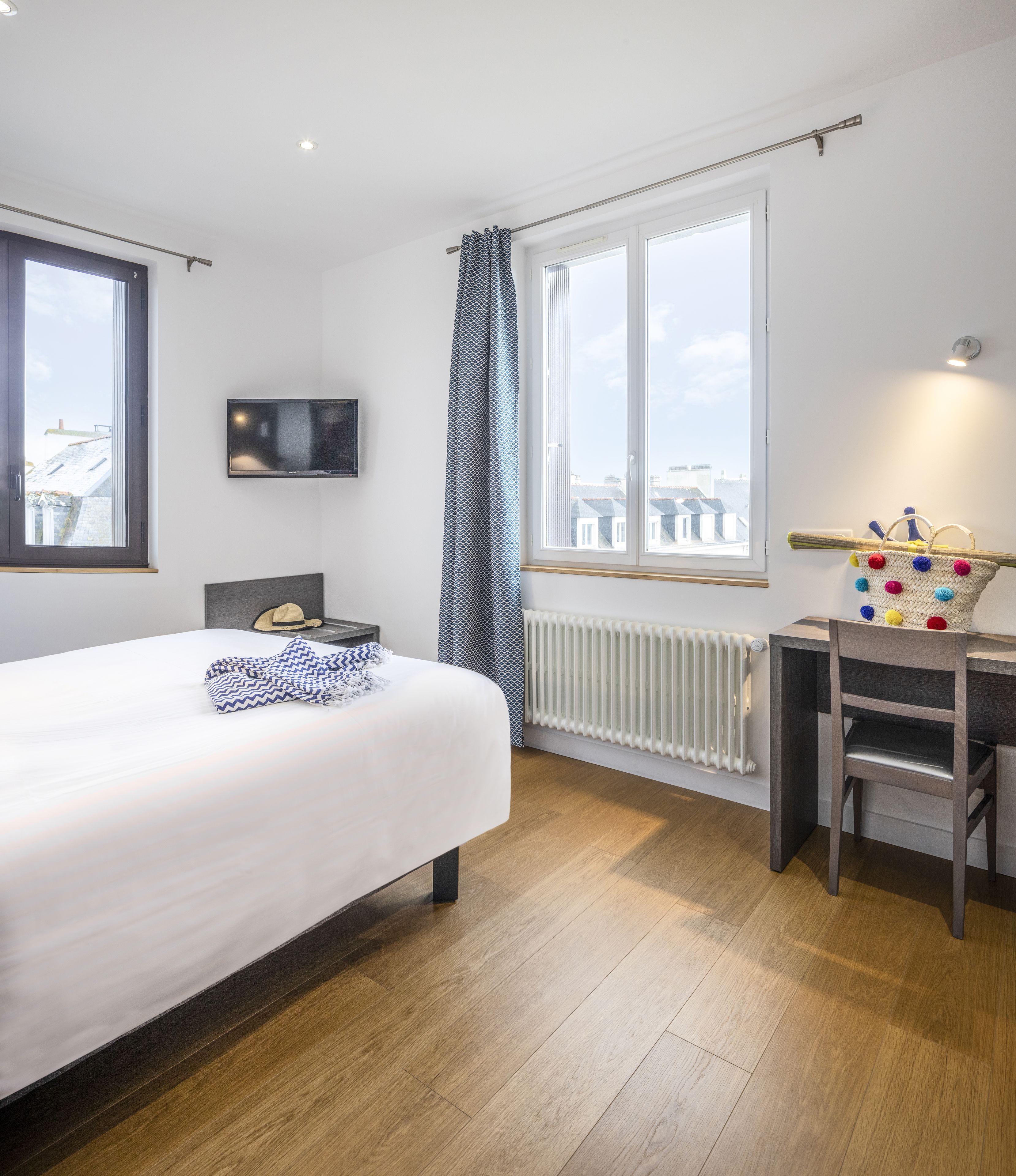 A modern bedroom with a large bed, a desk, a chair, a TV, and a window with a view of buildings.