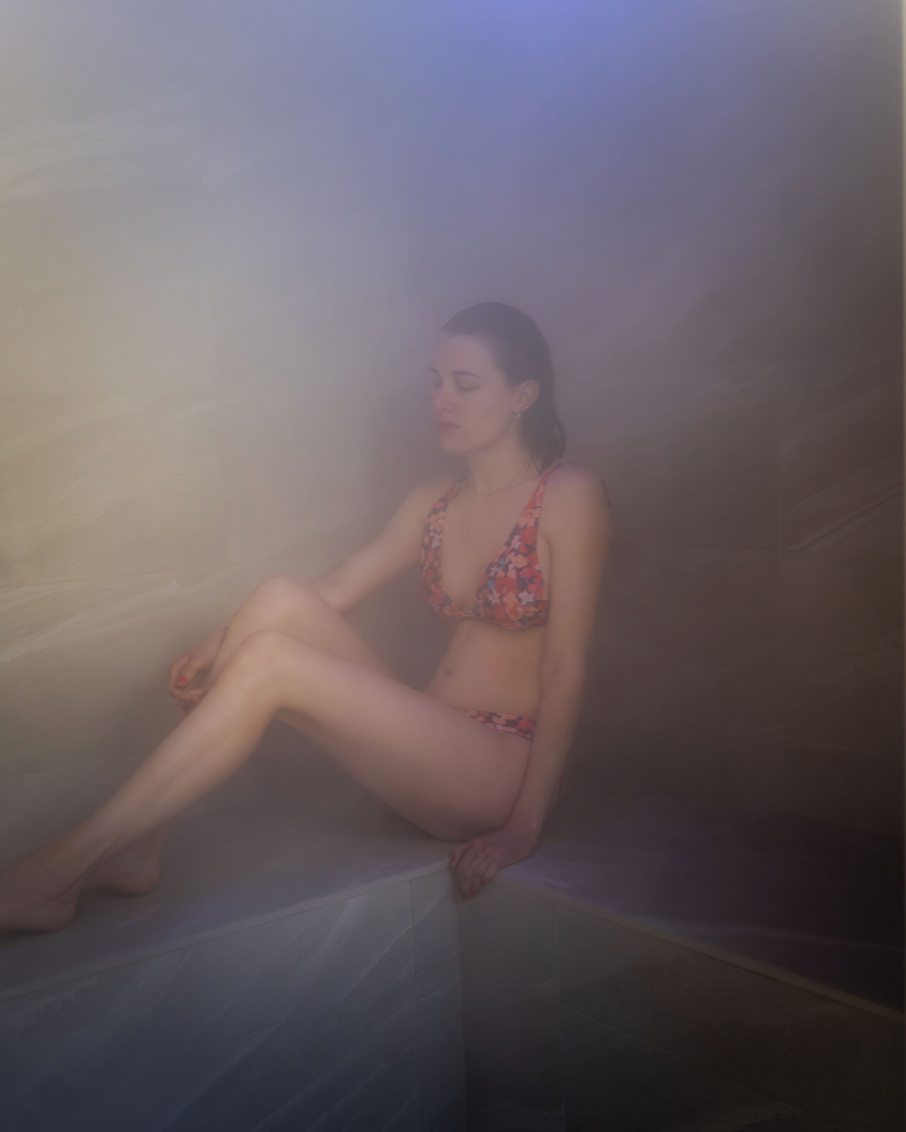 A person sitting in a steam room with a tiled floor and a plain wall.