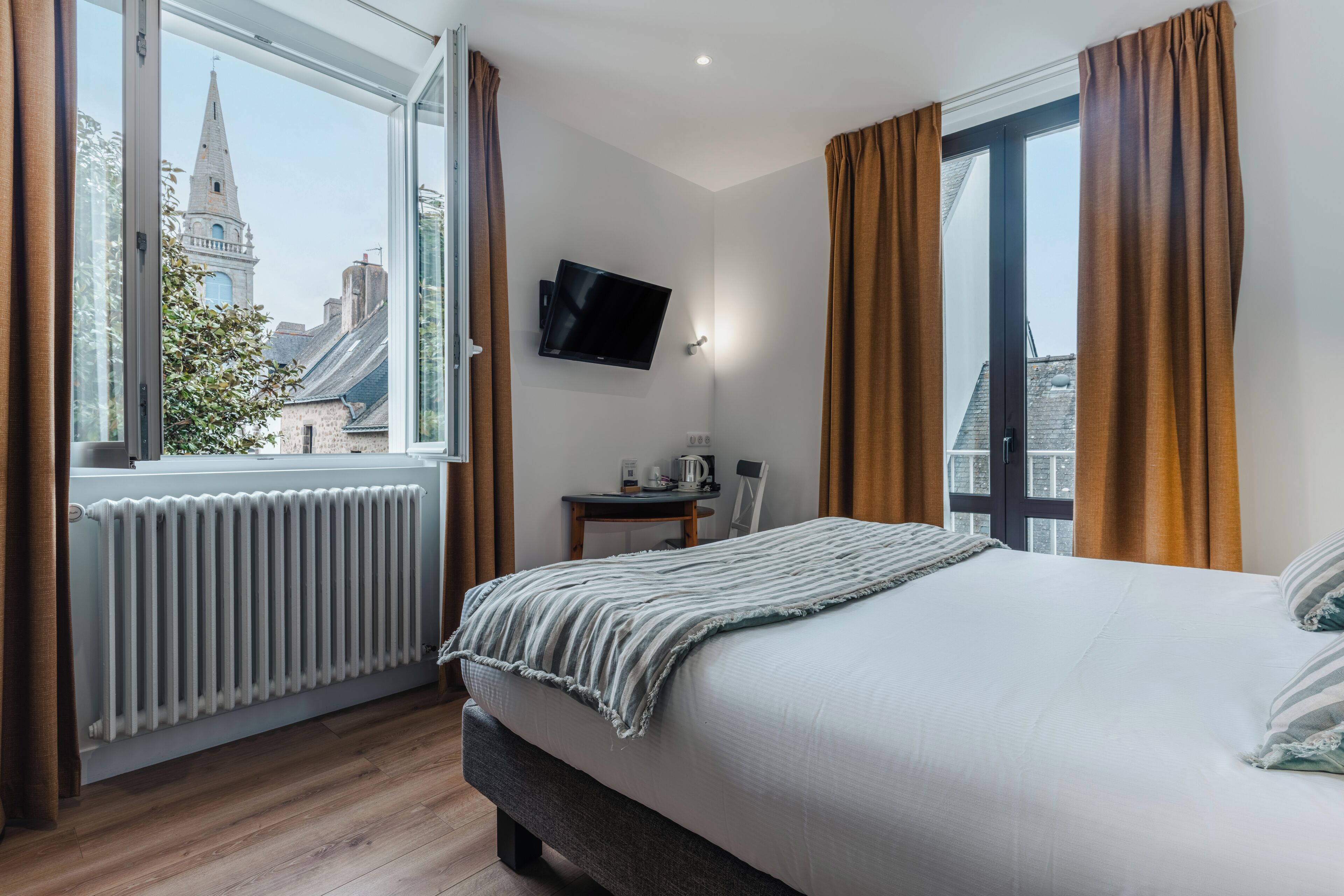 A hotel room with a large bed, a desk with a chair, a TV mounted on the wall, and a window with curtains.