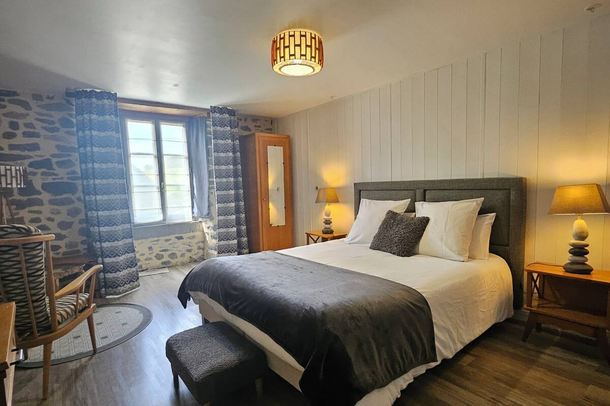 Een slaapkamer met een groot bed, een houten stoel, een klein tafeltje en een stenen muur.